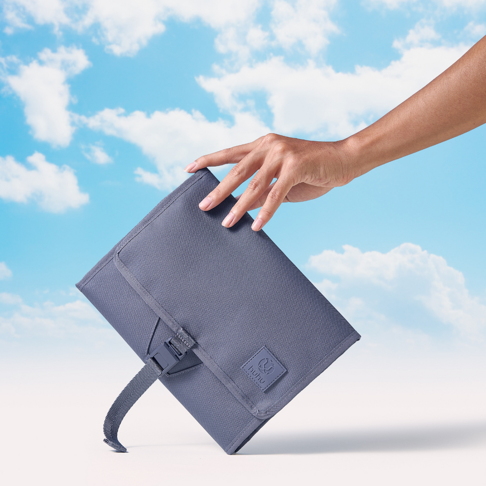 Hand holding blue changing wallet on angle against beige and blue background with white clouds.