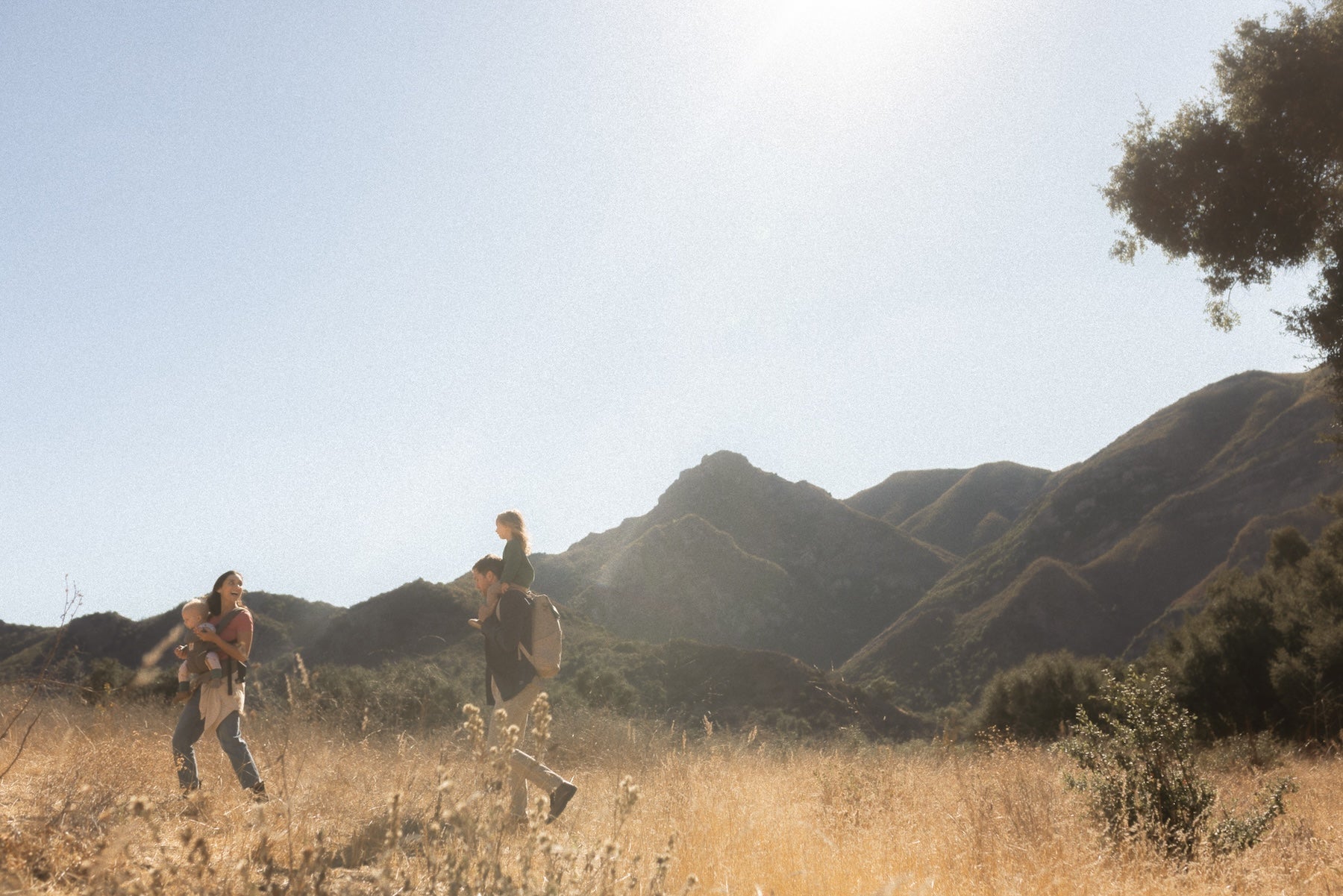 Mom wearing baby in carrier. Dad wearing beige backpack with toddler on shoulders. Walking in grassy field with mountains in the background.