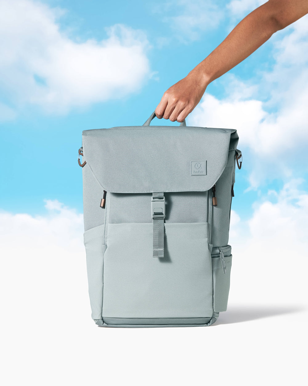 hand holding a green diaper backpack against a blue and white background with clouds