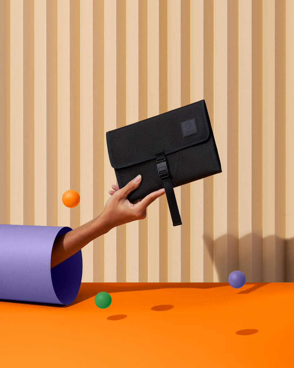 A hand holding a black wallet in front of a tan striped wall. Purple, green, and orange balls bouncing off of orange ground.